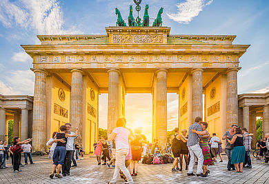 Gruppe glücklicher junger Menschen, die vor dem berühmten Brandenburger Tor tanzen, Symbol für Frieden und Einheit und historisches Wahrzeichen, in goldenem Abendlicht bei Sonnenuntergang mit blauem Himmel im Sommer, Berlin, Deutschland