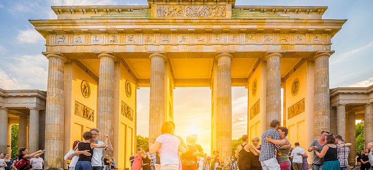 Gruppe glücklicher junger Menschen, die vor dem berühmten Brandenburger Tor tanzen, Symbol für Frieden und Einheit und historisches Wahrzeichen, in goldenem Abendlicht bei Sonnenuntergang mit blauem Himmel im Sommer, Berlin, Deutschland
