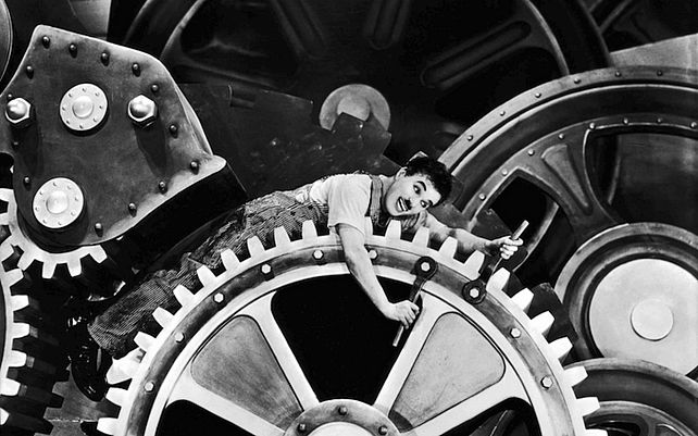Charlie Chaplin hängt zwischen riesigen Metallrädern fest. Bild schwarz/weiß